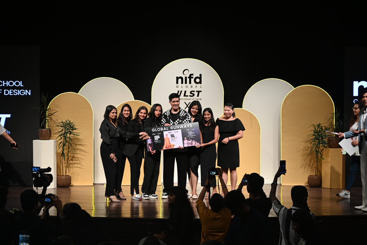 A group on stage holds a large certificate in front of a "nifd GLOBAL RUNWAYS" backdrop, as audience members capture the moment.