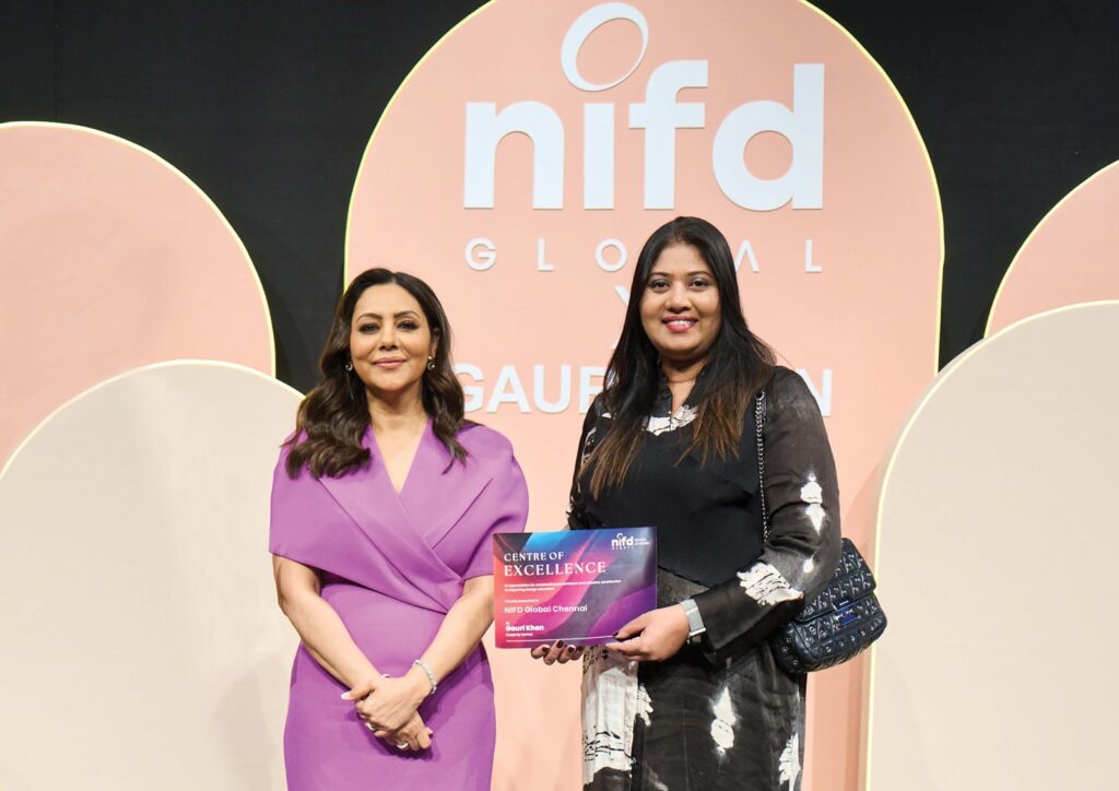 Two women stand before a "nifd Global" backdrop; one in a purple dress and the other in black and white holds a "Centre of Excellence" plaque.