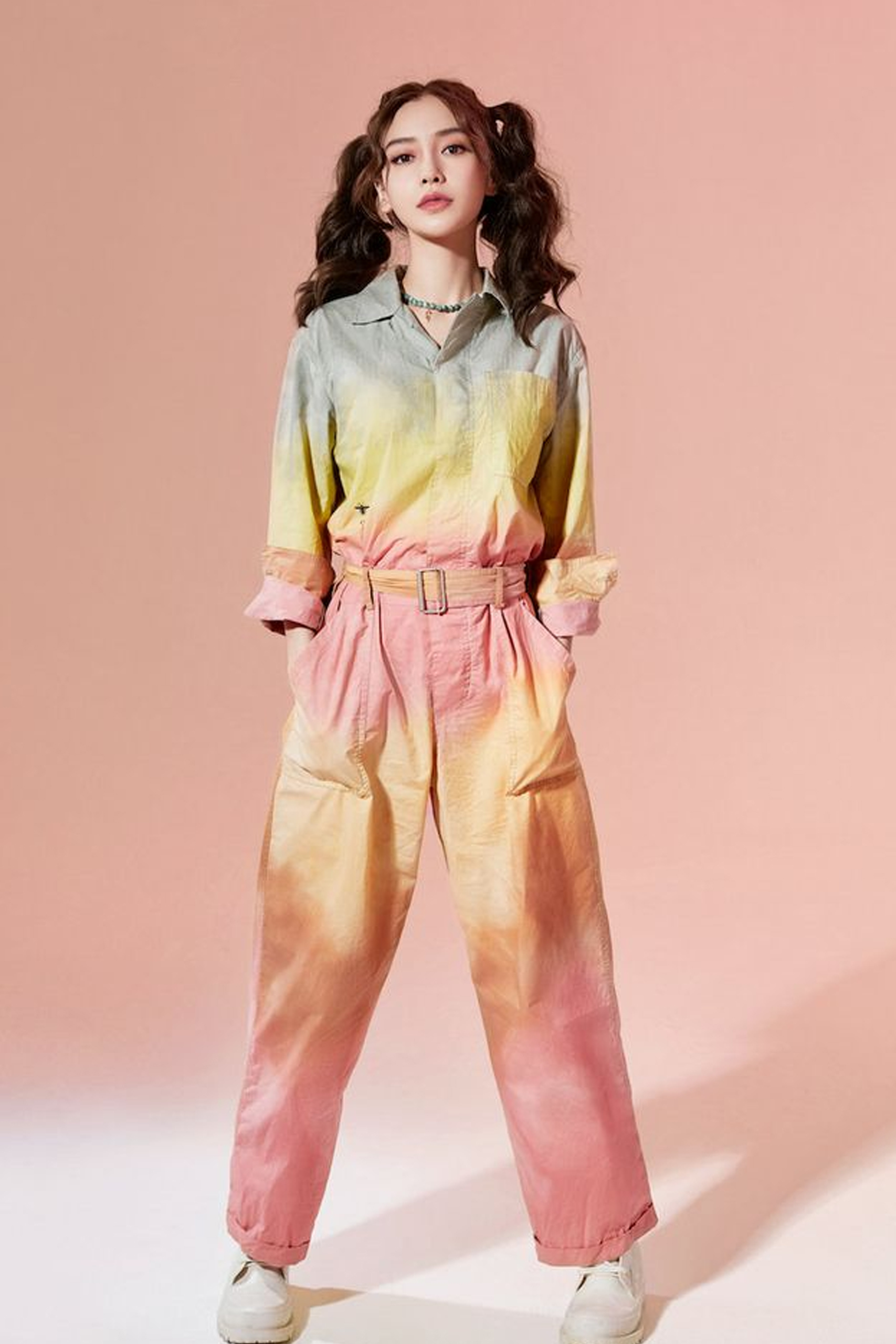 A confident young woman with wavy hair wears a pastel gradient outfit—green-to-yellow shirt and peach-to-pink pants—against a peach background.